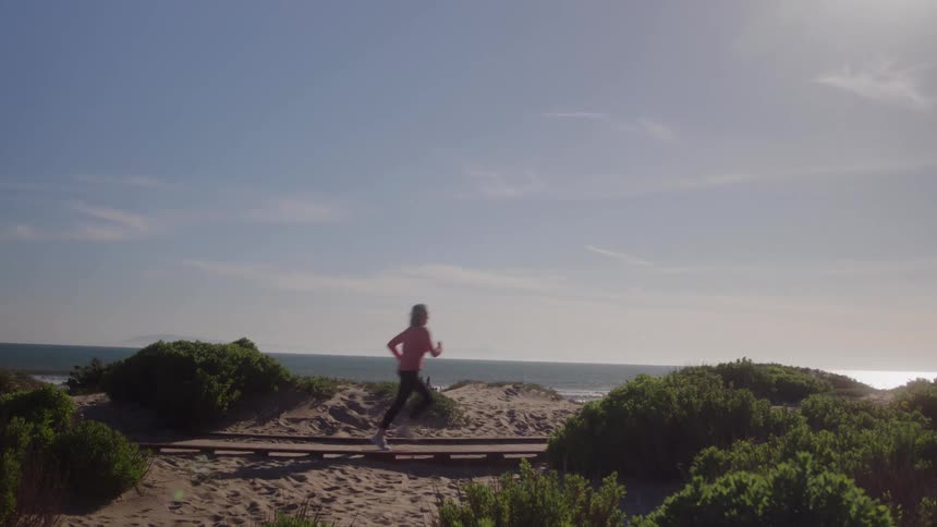 Woman running on a boardwalk through sand dunes under bright sunlight