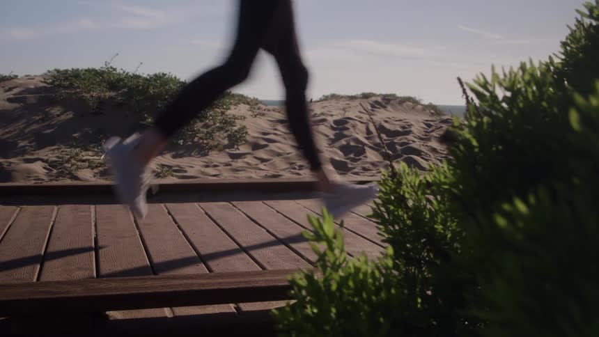A low angle close view shows a persons legs running across a wooden boardwalk that leads toward a sandy dune beach with the ocean visible in the distance and green foliage in the foreground