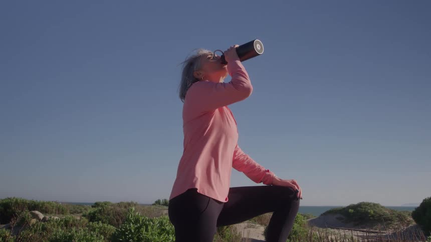 A woman in workout clothes standing outdoors drinking water from a thermos bottle