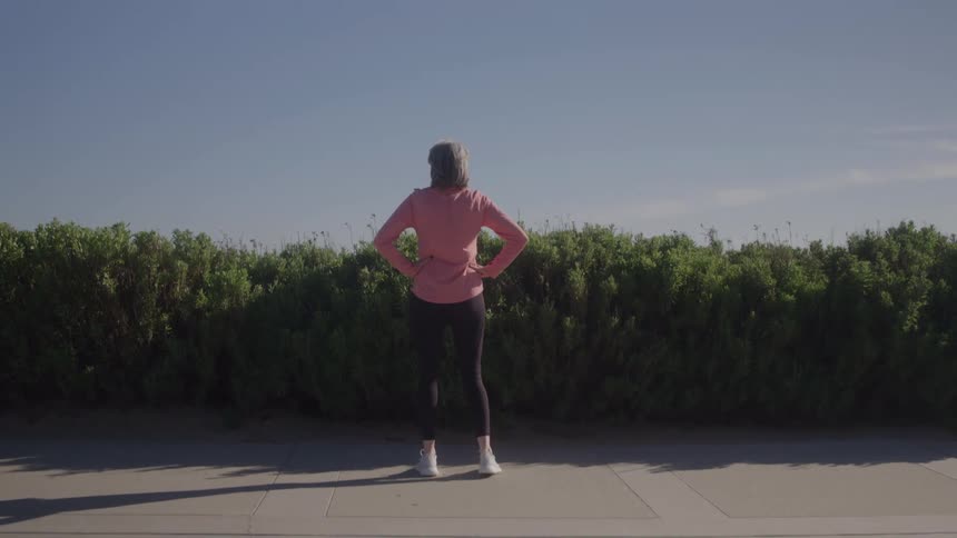 Person in pink top with hands on hips faces hedge under blue sky on sunlit pavement