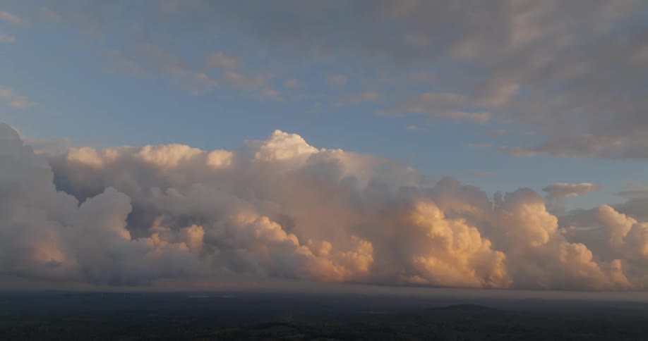 A beautiful wide view of the sky shows large towering cumulus clouds lit golden by the setting sun looming over dark hills below a pale blue sky
