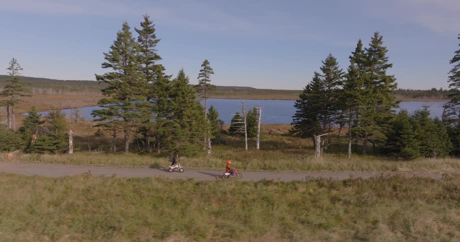 Two people ride bicycles on a road surrounded by pine trees with a lake in the background