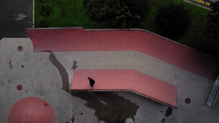 The video shows an overhead drone shot of a skate park with a skater carving back and forth on a section of redpainted ramp the ramp is part of a larger concrete park surrounded by green trees and foliage