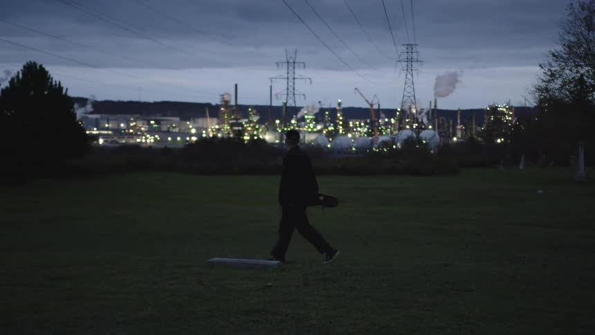 A man walks across a grassland at sundown with power lines stretching overhead and a distant power plant glowing with light