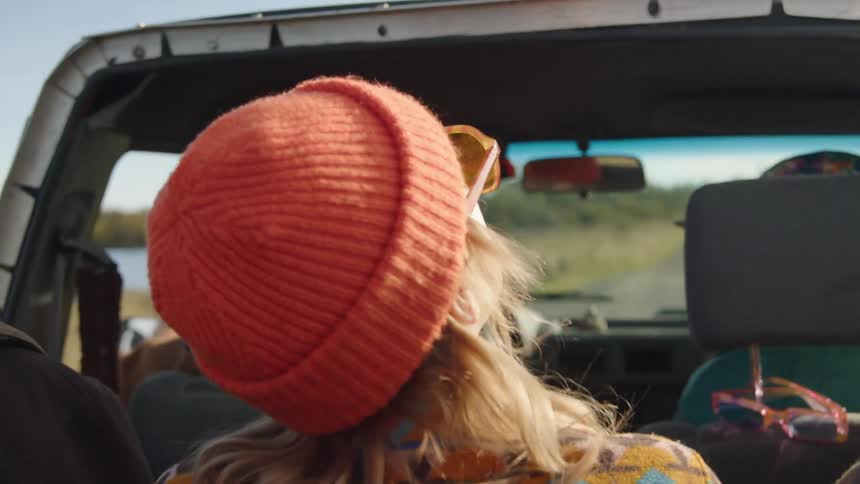 The video is an interior shot of a car focusing on a woman in the passenger seat wearing an orange knit beanie and reflective sunglasses she is looking away from the camera and the bright sun and trees of the road trip setting are visible throu
