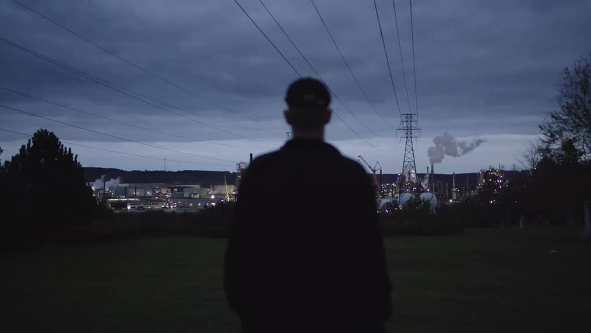 The video shows a silhouetted figure standing in a dark field facing a brightly lit industrial plant or refinery in the distance the scene is set at dusk or night with power lines and utility towers cutting across the cloudy sky