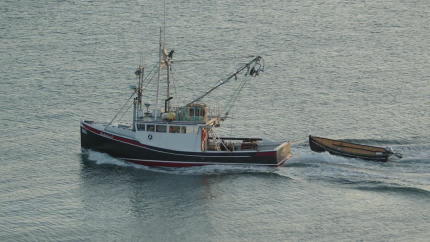 A classic red and black commercial fishing vessel speeds across the water pulling a small wooden dinghy behind it as the engine leaves a long white wake in the gray ocean