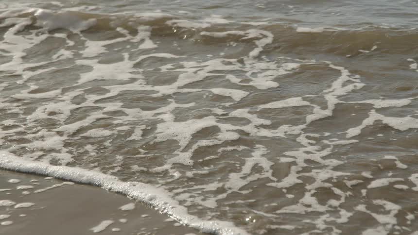 The video is an extreme closeup shot of waves gently breaking on a sandy shore the water is foamy and appears silverygray under a bright overcast sky giving the scene a soft muted look