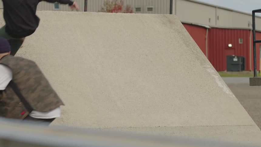 Two people consecutively ride skateboards up and down a slanted concrete wall in the foreground with an open paved area and industrial buildings visible in the background