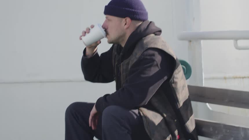 A man with a mustache sits outdoors in a dark vest holding and drinking from a white paper cup while looking offcamera in a hazy environment