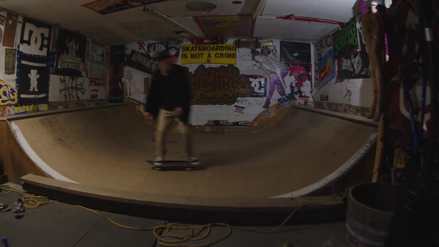A skateboarder rides across a wooden halfpipe ramp inside a garage whose walls are covered with posters and graffiti while another person stands by