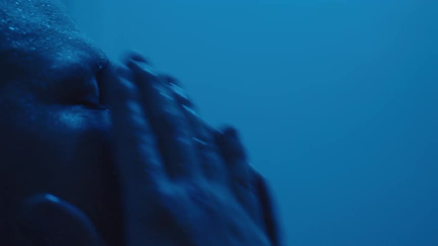 The video is an extreme closeup of a persons face partially submerged underwater the scene is bathed in a deep saturated blue light with water droplets clinging to the skin above the surface
