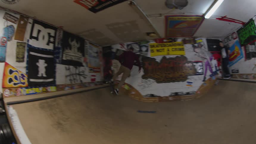 A skateboarder rides back and forth across a wooden miniramp inside a small room the walls of the space are covered in stickers and graffiti