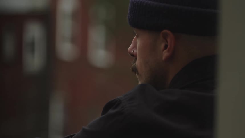 A man with a mustache wearing a black cap and jacket stands near a doorway his profile visible the background is out of focus
