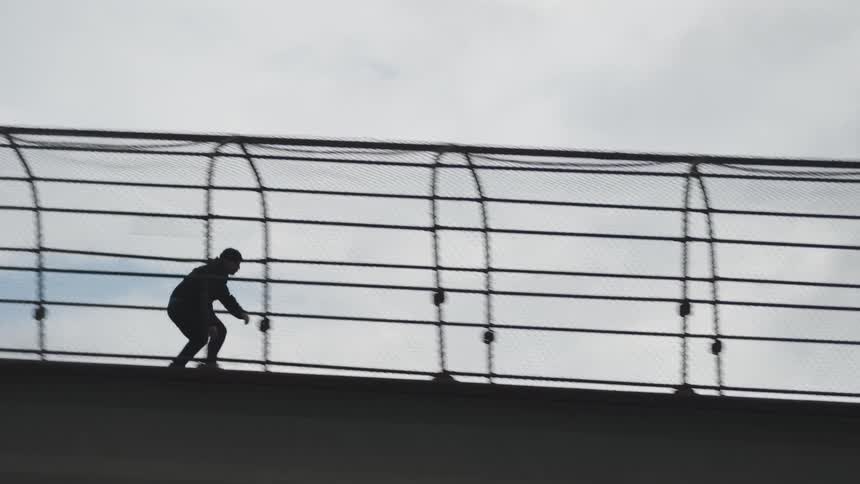 The video is an extreme sideprofile silhouette of a person walking across a caged pedestrian bridge or overpass against a bright sky the chainlink cage structure is prominent in the frame creating a grid pattern around the figure