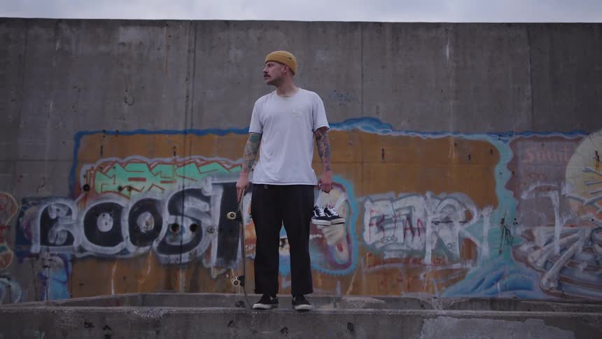 A man holding a skateboard stands by a graffiti covered wall outdoors