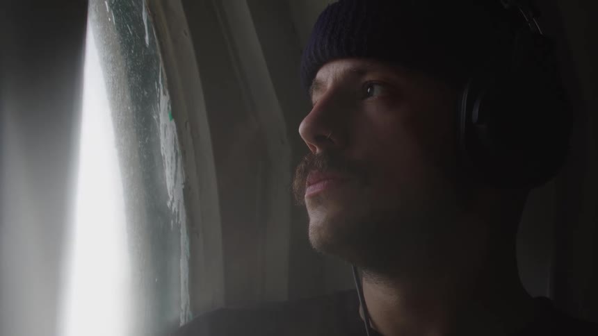 A camera shot shows a mans face in profile wearing headphones and a knit cap looking out a window framed by bright light while the rest of the setting remains in shadow