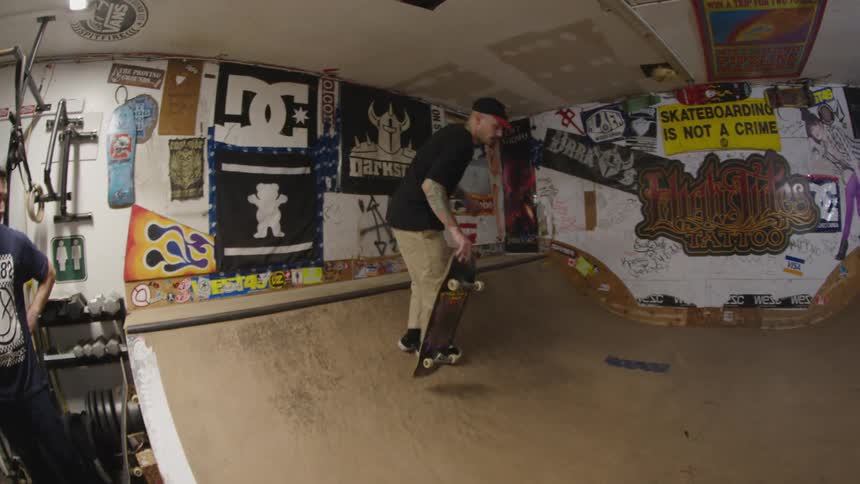 A person carrying a skateboard walks from a dark exterior into a brightly lit garage containing a skate ramp covered in stickers and art then jumps onto the board to start skating the curve