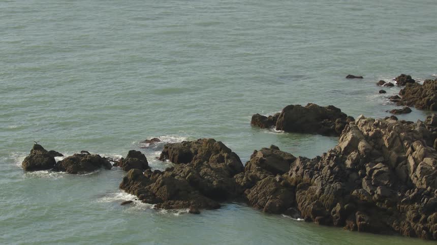 A high angle wide view shows jagged dark brown rocks partially submerged in the pale green water of the ocean under a clear bright sky