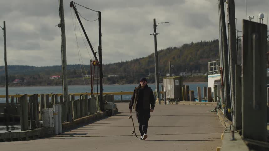 A person wearing dark clothing and a cap walks toward the camera along the concrete surface of a long open pier holds a skateboard at their side and is framed against a background of water and wooded hills under a cloudy sky