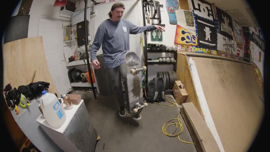Person performs skateboard trick midair in indoor space decorated with decks tools and posters