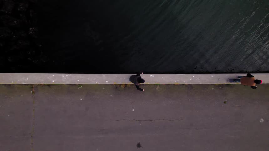 An overhead aerial view shows two men walking along a stark gray concrete pier next to dark water