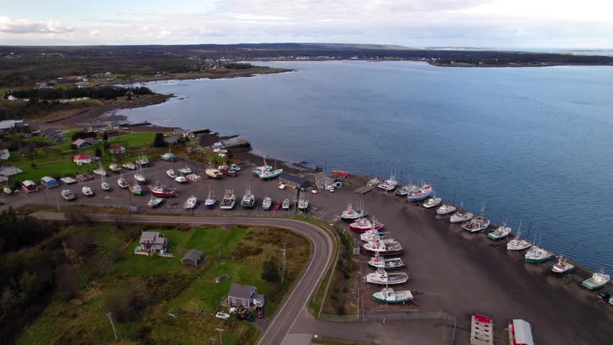 An aerial view shows a small seaside town with dozens of fishing boats pulled ashore at a dry dock next to a large blue bay under a partially cloudy sky