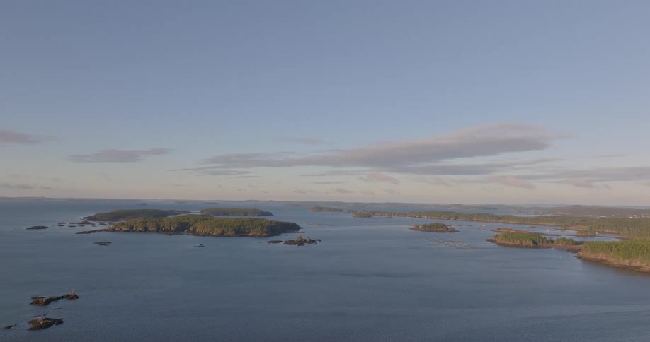 An aerial wide shot shows a calm body of water dotted with numerous dark treecovered islands and a wooded mainland coast under a clear light blue sky