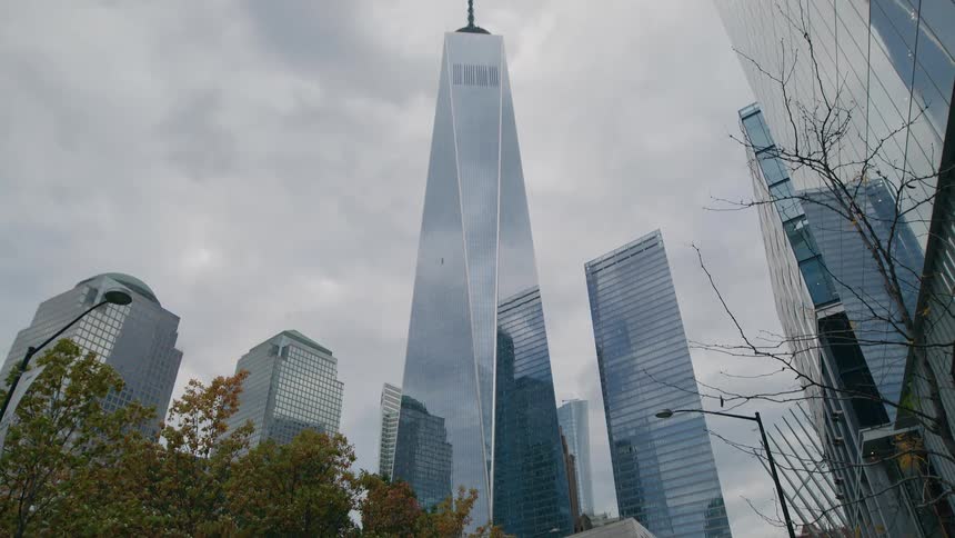 A low angle wide view shows the one world trade center skyscraper surrounded by other modern glass buildings and fall foliage against a cloudy pale sky in new york city