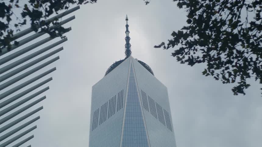 The upper section and antenna of the one world trade center skyscraper framed by blurred trees and a modern building against an overcast sky