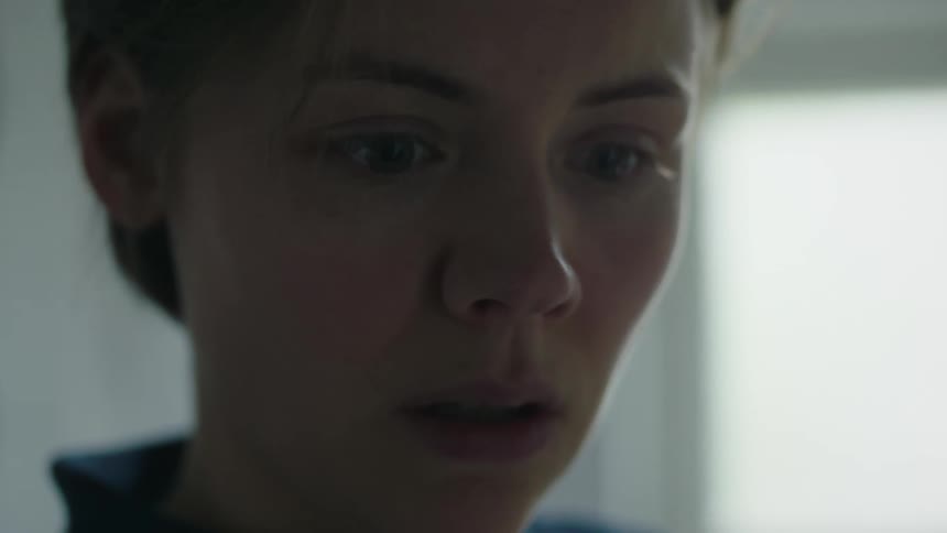 A womans face is shown in closeup her eyes wide and gazing downward with an expression of distress before she slowly bows her head and turns her profile away from the camera