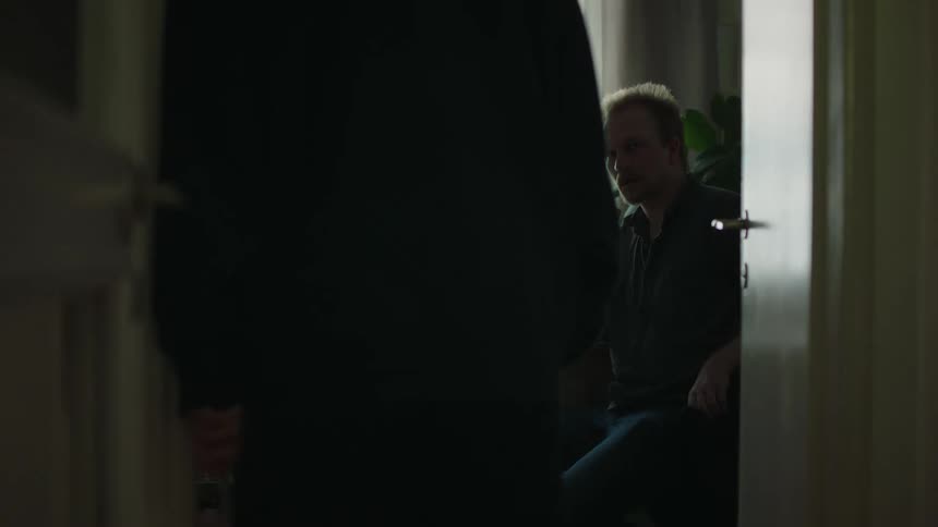 A man sits alone at a table near a brightly lit window his figure silhouetted and head lowered as he contemplates while a second person walks quickly through the darkened doorway in the foreground