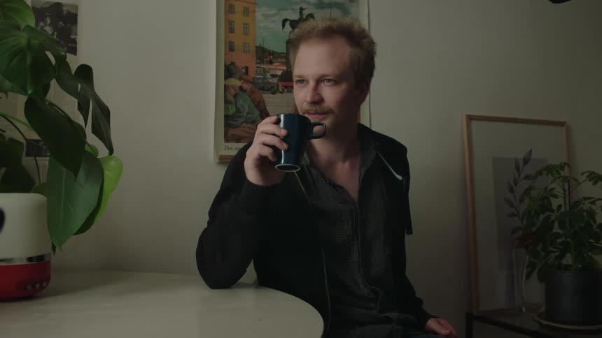 A man sits at an indoor table while a second person pours liquid from a carafe into his dark mug which he then raises to his mouth to drink