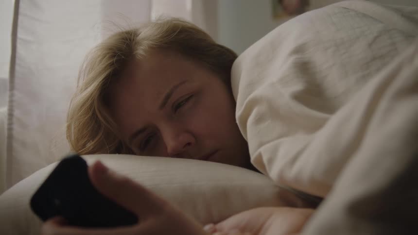 Woman lying in lightcolored bed holds phone while resting with halfclosed eyes under soft light