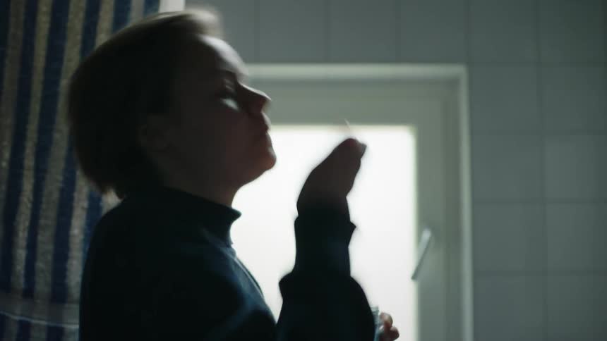 A profile view of a woman with short blonde hair wearing a dark turtleneck sweater as she takes a pill while looking in a bathroom mirror with bright light coming from a window
