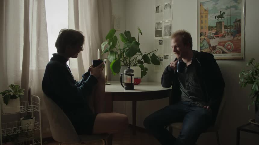 Man and a woman with a cup sit at a round table surrounded by plants in a bright room