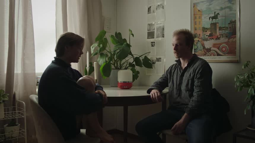 A man sitting on a chair and a young woman enters the room and sits on another chair she looks tense and they start talking