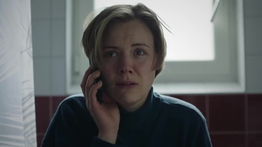 A close view shows a distressed woman with short blonde hair wearing a dark turtleneck as she holds a cell phone to her ear with an intensely worried expression in a softly lit bathroom