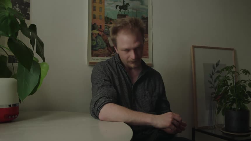 A man sits at a white table leans an arm on the table surface looks away from the camera and down and the background contains household decorations and potted greenery