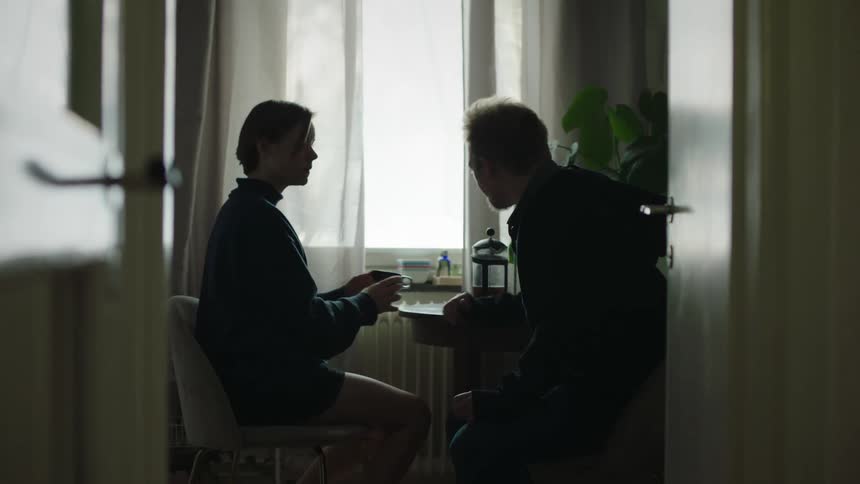The video shows a medium shot of two people sitting indoors at a table near a bright window viewed through a doorway the woman is facing the man holding a mug while the man is silhouetted and leaning forward in conversation