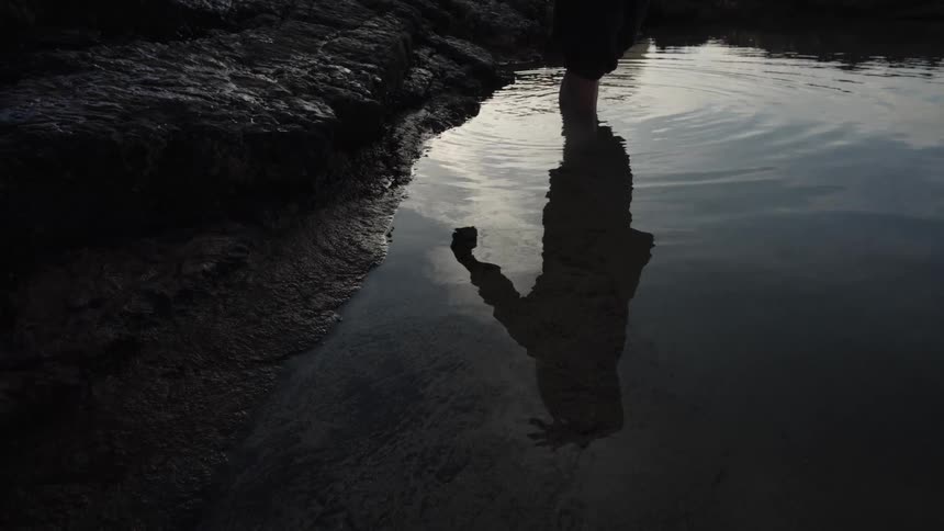 A low angle shot captures a person with bare legs standing in shallow water next to dark rocks their silhouette and reflection stretching across the calm surface of the water