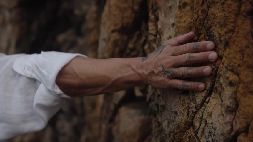 A closeup shot focuses on a persons tattooed hand bracing against a reddishbrown textured rock face the person is wearing a longsleeved white shirt as they hold onto the natural surface