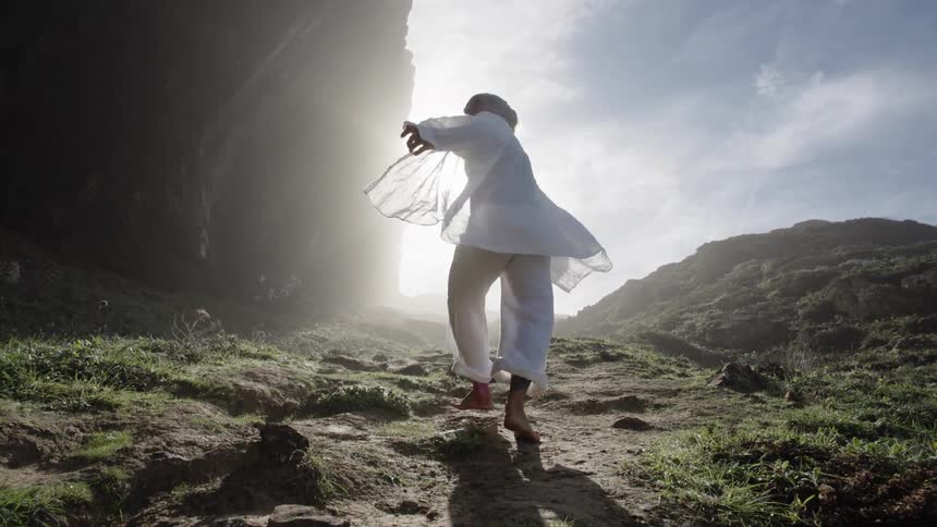 Person in ceremonial attire walks barefoot over rocky terrain toward light between mountains