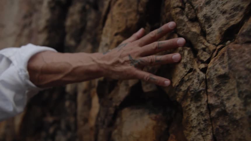 A close view of a persons tattooed hand and arm wearing a white shirt pressed flat against the rough dark brown and orange surface of a rock face