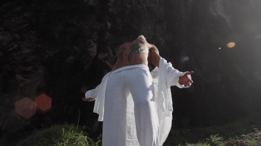 A grayhaired bearded man wearing a white shirt and trousers leans backward while standing on soil his shirt marked with a tattoo under the bright sun