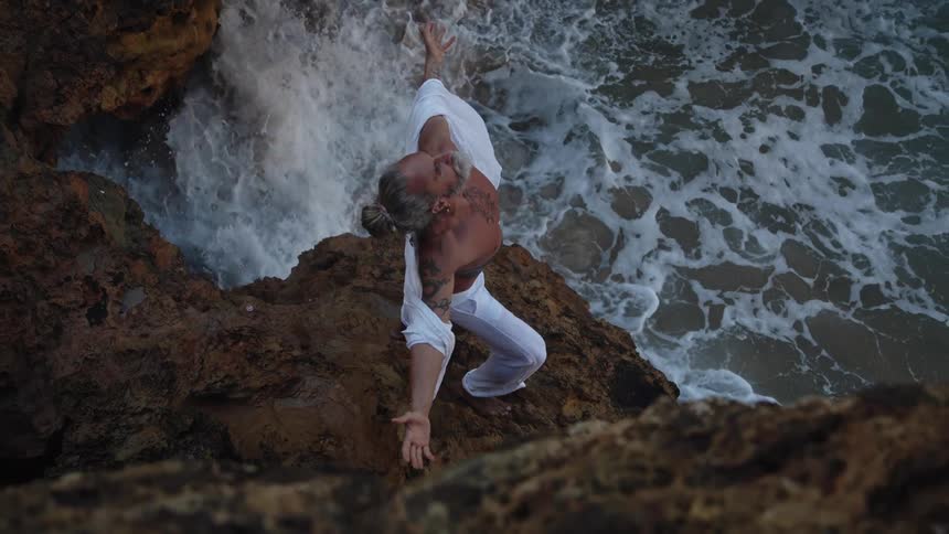 A middleaged man with gray hair and a beard stands shirtless on a cliff by the shoreline at midday his white shirt fallen below his shoulders and white trousers on arms spread wide as ocean waves crash against the rocks below