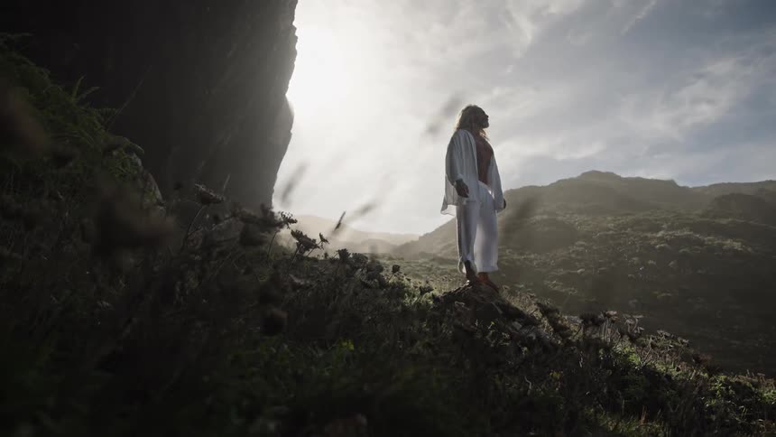 Man in white robe with dark sash standing on hillside lit by sun through clouds