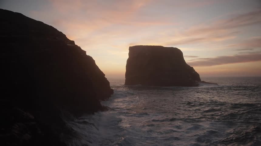A long view of a dark sea stack silhouette against a dramatic sunset sky with choppy ocean waves crashing against the rocky foreground cliff