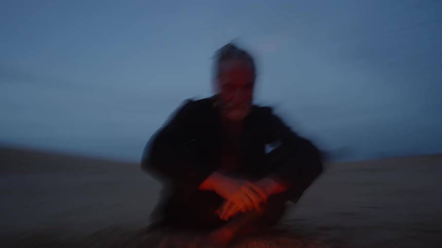 An elderly man wearing dark clothing and with hair tied up sits on the ground next to a campfire at dusk in an open dark environment with the flames casting an orange light