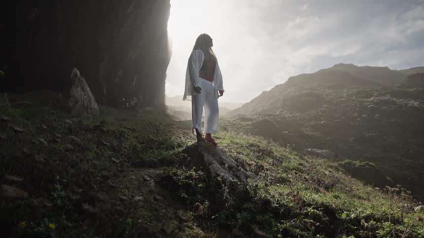 The video shows a man standing on a rocky grassy mountain trail looking up into a bright sundrenched sky he is wearing a white open shirt and is silhouetted against the bright light creating a strong contrast with the dark green and brown